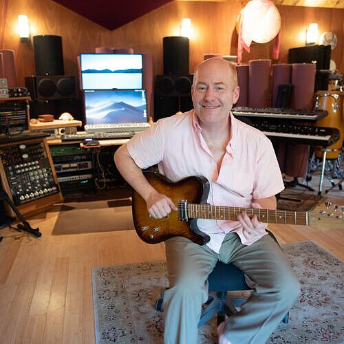 a very handsome man sits in a chair with a grogeous t shaped guitar, the walls of the room lit warmly - clearly a place in which fabulous music is made!